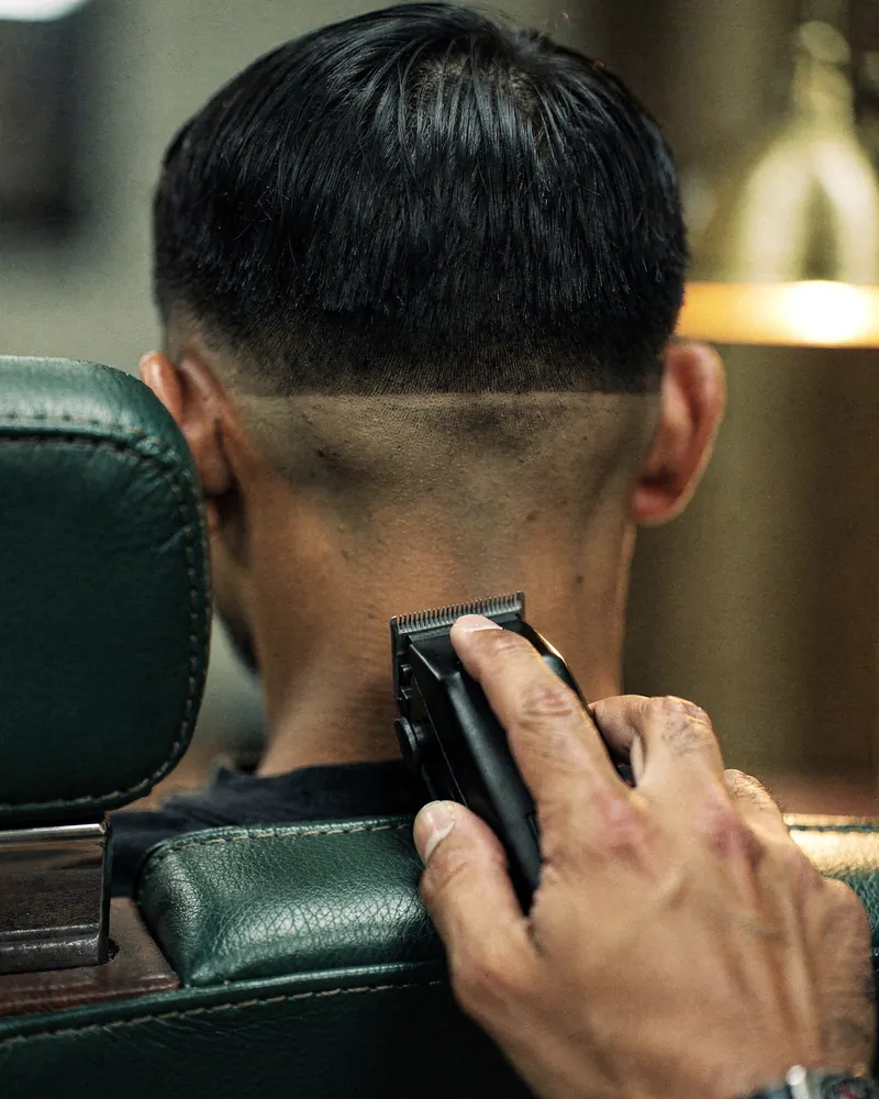 A barber's hand runs clippers along a client's nape to finish a fade, seen from behind in a green leather chair.